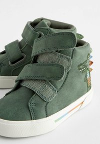 Zelené vysoké tenisky vyrobené z texturovaného syntetického materiálu s suchým zipem, ozdobené barevnými pruhovanými detaily na podrážce a bočním designu.