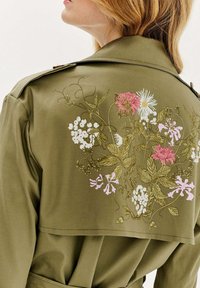 Oliwkowy trench coat z floralnym haftem w kolorach różowym, białym i żółtym na plecach, z wyraźnie wyprofilowanym kołnierzem i widocznymi detalami szwu.