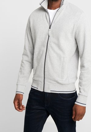 Mann trägt eine hellgraue Reißverschlussjacke mit gerippten Bündchen und Saum, kombiniert mit dunkelblauen Jeans, steht vor einem schlichten Hintergrund.