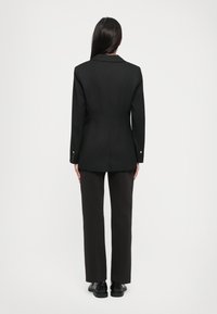 Svart blazer med strukturerte skuldre, enkelt bakventil og gullknappeakenter, kombinert med rettkuttede mørke bukser og svarte sko.