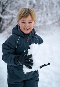 Egy gyermek tengerészkék télikabátban egy nagy hógolyót tart a kezében. A kabát kapucnival és fehér varrással díszített. Fekete kesztyűk. Havas háttér.