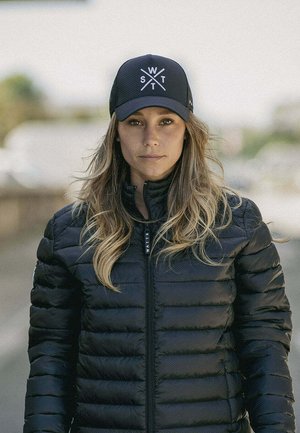 Vrouw met lang haar, gekleed in een zwart doorgestikt jack en een zwarte pet met witte letters W, S, T, staat buiten en kijkt vooruit.