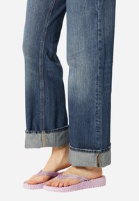 Blauwe denim jeans met omslagen, gedragen met roze gevlochten teenslippers op blote voeten tegen een witte achtergrond.