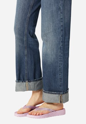 Blauwe denim jeans met omslagen, gedragen met roze gevlochten teenslippers op blote voeten tegen een witte achtergrond.