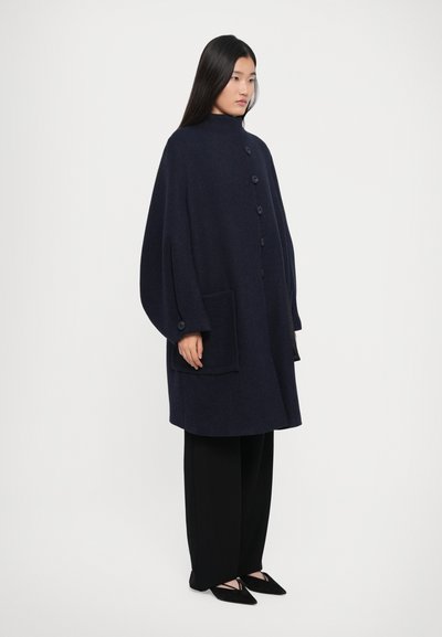 Femeie îmbrăcată cu un palton cu nasturi oversized de culoare bleumarin, având buzunare mari, pantaloni negri evazați și balerini negri cu curea.