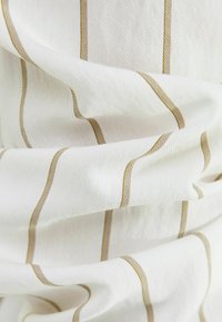 Bílá látka s texturovanou vazbou, se štíhlými taupe pruhy probíhajícími vertically. Materiál vypadá jemně a mírně pomačkaně.