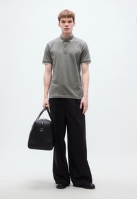 Γκρι πόλο πουκάμισο με αντίθετη λεπτομέρεια, σε συνδυασμό με φαρδιά μαύρη παντελόνια. Το μοντέλο κρατά μια μαύρη τσάντα με λεία υφή και στρογγυλεμένο σχήμα.