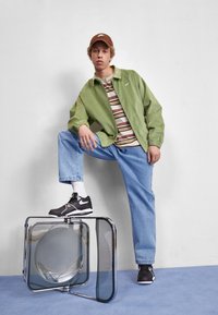 Grön nylonjacka, randig t-shirt, ljusblå jeans och svarta sneakers med vita accenter. Modellen poserar på en glasstruktur.