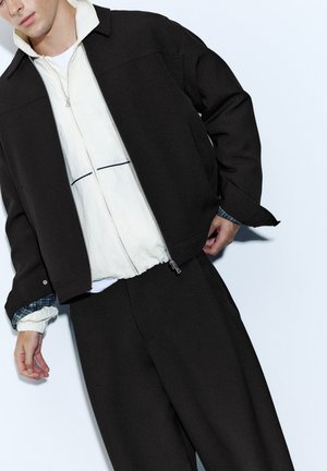 Homme portant une veste et un pantalon noirs sur un sweat à capuche blanc zippé avec des manches de chemise à carreaux bleus visibles sur fond blanc.