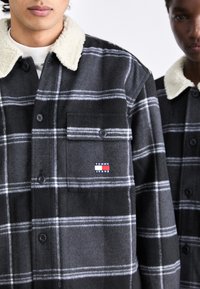 Karierter Flanellhemd in Schwarz und Grau mit weißen Streifen, ausgestattet mit einem Schalkragen aus Schafswolle, Knopfleiste und einer Brusttasche mit Logodetail.