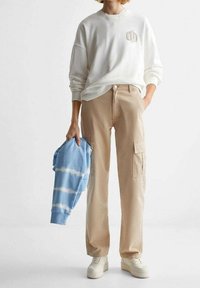 Světle šedá mikina s textem "ALWAYS", béžové cargo kalhoty s bočními kapsami, drží modrou tie-dye mikinu a má na sobě bílé tenisky.