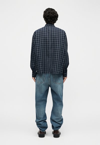 Άτομο με σγουρά μαλλιά που φοράει μπλε-ναυτικό καρό πουκάμισο και άνετο ξεθωριασμένο μπλε τζιν, στέκεται με την πλάτη σε λευκό φόντο.