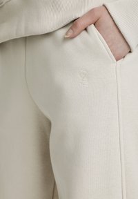 Pantaloni sportivi beige realizzati in morbido misto cotone, con tasche laterali e un piccolo logo ricamato sull'anca. Tessuto liscio e vestibilità rilassata.