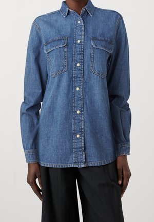 Person trägt ein langärmliges blaues Denim-Hemd mit zwei Brusttaschen und schwarzen Hosen vor neutralem Hintergrund.
