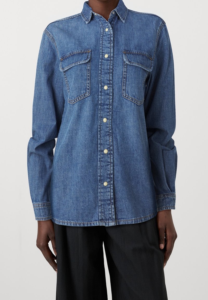 Personne portant une chemise en denim bleu à manches longues avec deux poches poitrine et un pantalon noir sur un fond uni.
