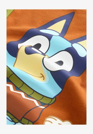 T-shirt in cotone con il volto di un cane cartoon in blu, giallo e blu scuro, su uno sfondo arancione e con dettagli di un maglione verde.