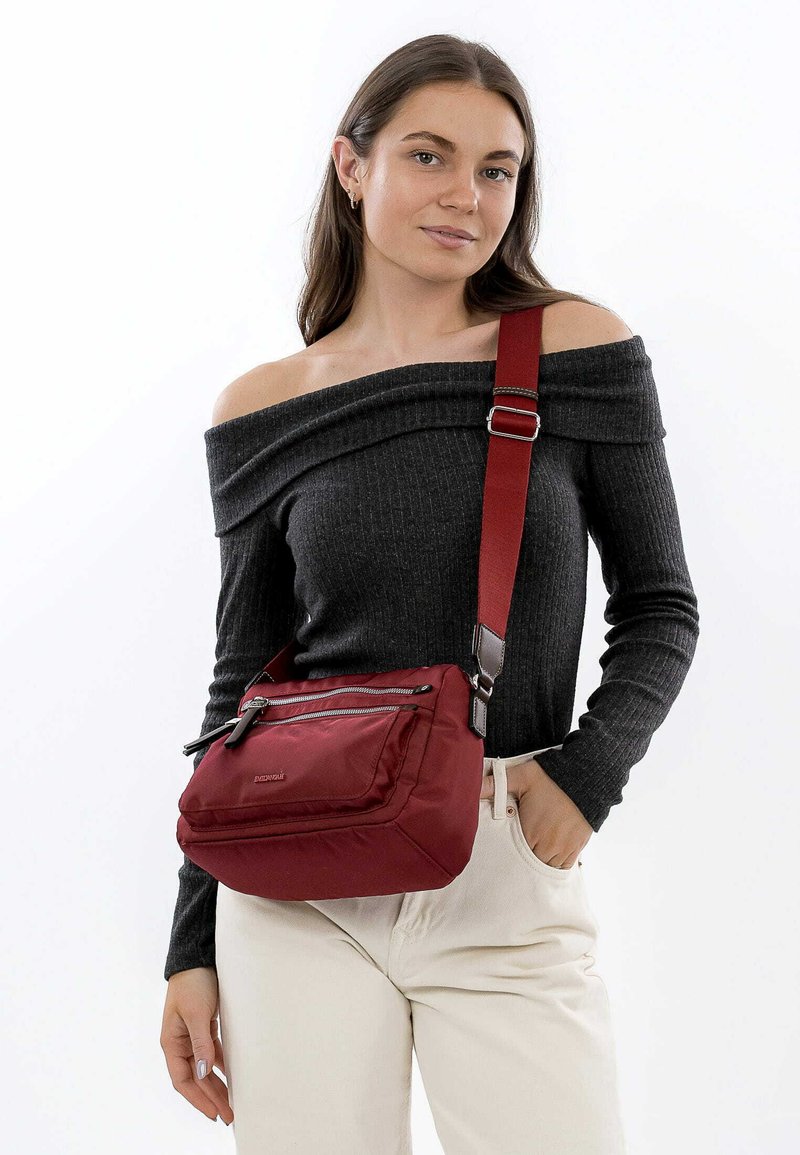 Červená nylonová crossbody taška s přední kapsou na zip, nastavitelným popruhem a měkkou texturou. Modelka má na sobě šedý top bez ramínek a béžové kalhoty.