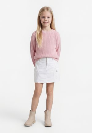 Jong meisje staand met handen achter de rug, gekleed in een roze trui, witte rok met zakken en beige enkellaarzen op een witte achtergrond.