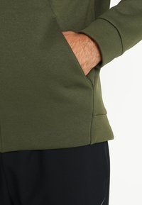 Sudadera de color verde oliva con una textura suave, que presenta un bolsillo frontal y puños acanalados. Combinada con pantalones negros, resaltando el contraste de color.