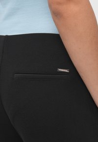 Τα μαύρα παντελόνια διαθέτουν μια πίσω τσέπη με διακριτική ασημένια λεπτομέρεια. Το ύφασμα έχει λεία υφή και ραμμένη κοπή.