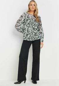 Blusa a fantasia bianco e nero con maniche lunghe trasparenti, scollatura rotonda, abbinata a pantaloni neri a gamba larga e scarpe con tacco nere.
