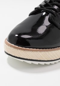 Chaussure en cuir verni noir à bout arrondi, dotée d'une semelle intermédiaire en corde beige texturée et d'une semelle extérieure en caoutchouc blanc. Des lacets noirs complètent le design.