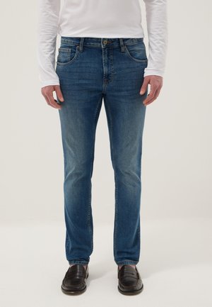 Man wearing blue slim-fit jeans, white long-sleeve shirt, and dark brown leather loafers standing against a plain background.