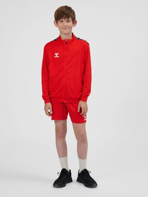Junge steht in roter Sportjacke und Shorts mit weißen Crew-Socken und schwarzen Turnschuhen vor neutralem Hintergrund.