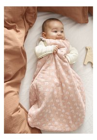 Saco de dormir acolchado en color rosa con un patrón de arcoíris blanco, tejido suave, colocado sobre ropa de cama blanca con cojines de color óxido y un juguete de madera cerca.