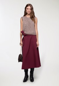 Beżowy sweter bez rękawów z wzorem w warkocze, w połączeniu z bordową spódnicą maxi z bocznym detalem w postaci kokardy. Czarne botki na obcasie dopełniają całość.