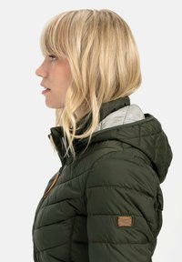 Oliven grøn pufferjakke med lynlås og hætte. Har en quiltet tekstur og en lysegrå foring, samt et lille logo-patch.