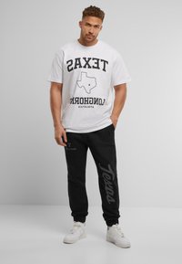 Bijela majica s natpisom "TEXAS" i nacrtanom siluetom Texasa, u kombinaciji s crnim trenirkama koje s jedne strane imaju veliki natpis "Texas". Bijele tenisice.