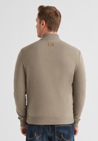 Hellbrauner Pullover mit hohem Kragen, gerippten Bündchen und Saum, mit einem Lederpatch am hinteren Kragen. Weiches, strukturiertes Material.
