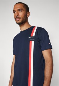 Camiseta de algodón azul marino con un diseño de rayas en blanco y rojo, logo "Champion" en el pecho y mangas cortas con acentos de logo en los puños.