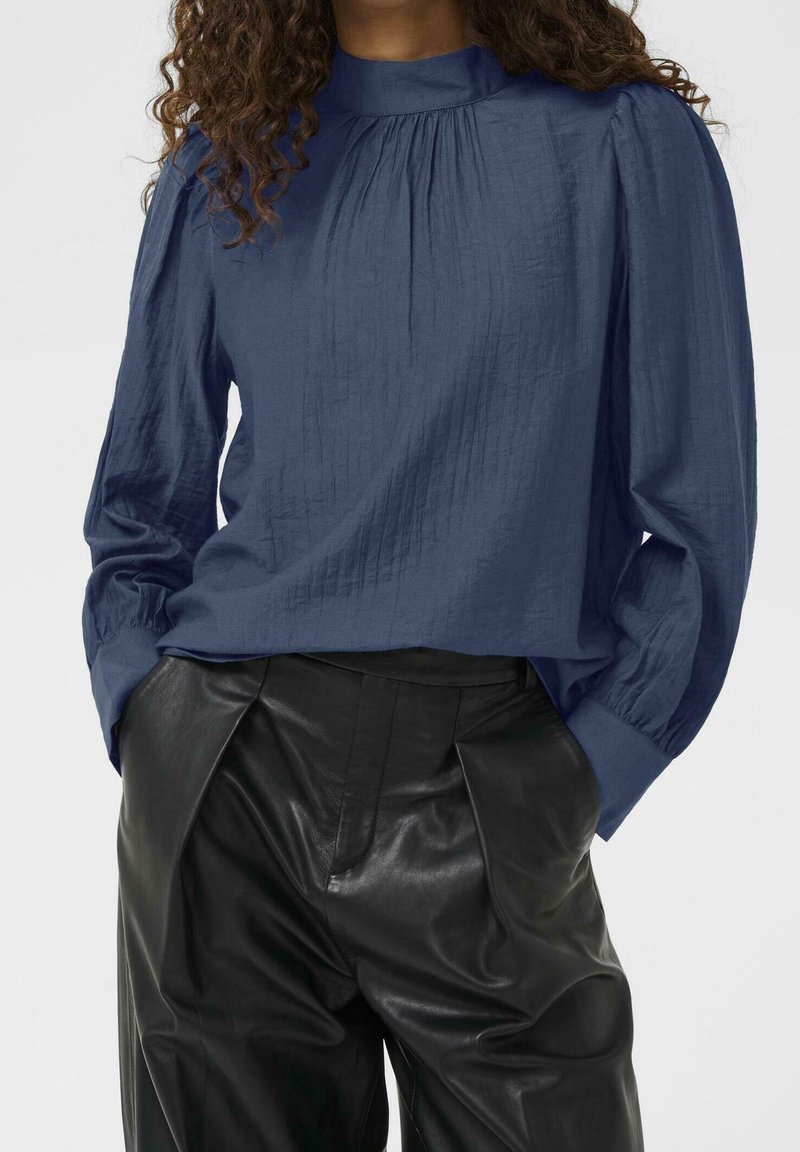 Femme portant un chemisier bleu marine à manches longues avec des détails froncés et un pantalon en cuir noir taille haute, mains dans les poches.