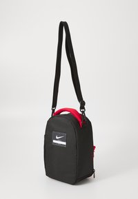 Svart nylonveske med rund form, rød topphåndtak, justerbar svart stropp og et logo-merke foran. Har en glidelåslomme på siden.