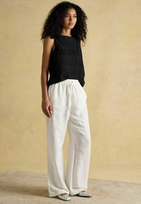 Melnā bez piedurknēm topiņš ar teksturētu rakstu, saskaņots ar brīvām balta linu biksēm un sudrabainām kurpēm. Mīksts, dabisks auduma apdare.