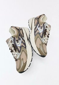 Scarpe sportive beige e marroni con tomaie in mesh, colli imbottiti, suole testurizzate e accenti bianchi a contrasto. Presentano lacci per una calzata sicura.