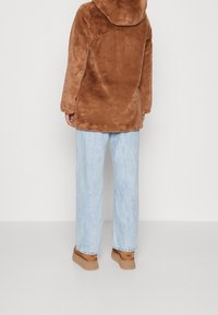 Brauner Kunstpelzmantel mit Kapuze, legerer Schnitt, getragen über hellblauen Jeans mit weitem Bein und braunen Schuhen. Glatte Textur und weiches Aussehen.