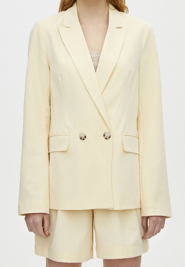 Femme portant un blazer croisé crème à deux boutons et un short assorti, debout devant un fond uni.