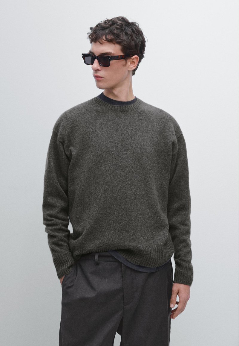 Maglione pullover in lana grigio scuro con collo e polsini a coste, abbinato a pantaloni scuri. Indossato con occhiali da sole rettangolari neri.
