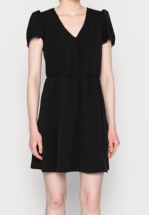 Kvinde iført en kort sort kjole med puffede korte ærmer og V-hals, stående mod en ensfarvet lys baggrund.