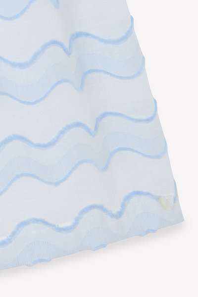 Λευκό ύφασμα με μπλε κυματιστό μοτίβο· διαθέτει μια υφή με αφράτο, ανοιχτό μπλε περίγραμμα και ένα μικρό διακοσμητικό σε σχήμα καρδιάς στο τελείωμα.