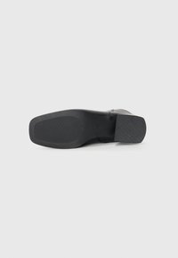 Zapato de tacón negro con parte superior de cuero liso, suela de goma texturizada y tacón cuadrado. La base tiene un patrón estriado para mayor tracción.