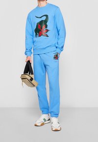 Man wearing light blue sweatshirt and sweatpants with green crocodile and red flower designs, holding a beige and black bag.