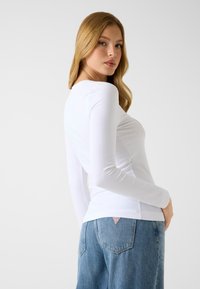 Langärmeliges weißes Oberteil mit figurbetontem Schnitt, weicher Textur und rundem Ausschnitt. Kombiniert mit hellblauen High-Waist-Denim-Jeans mit einem Logoetikett.