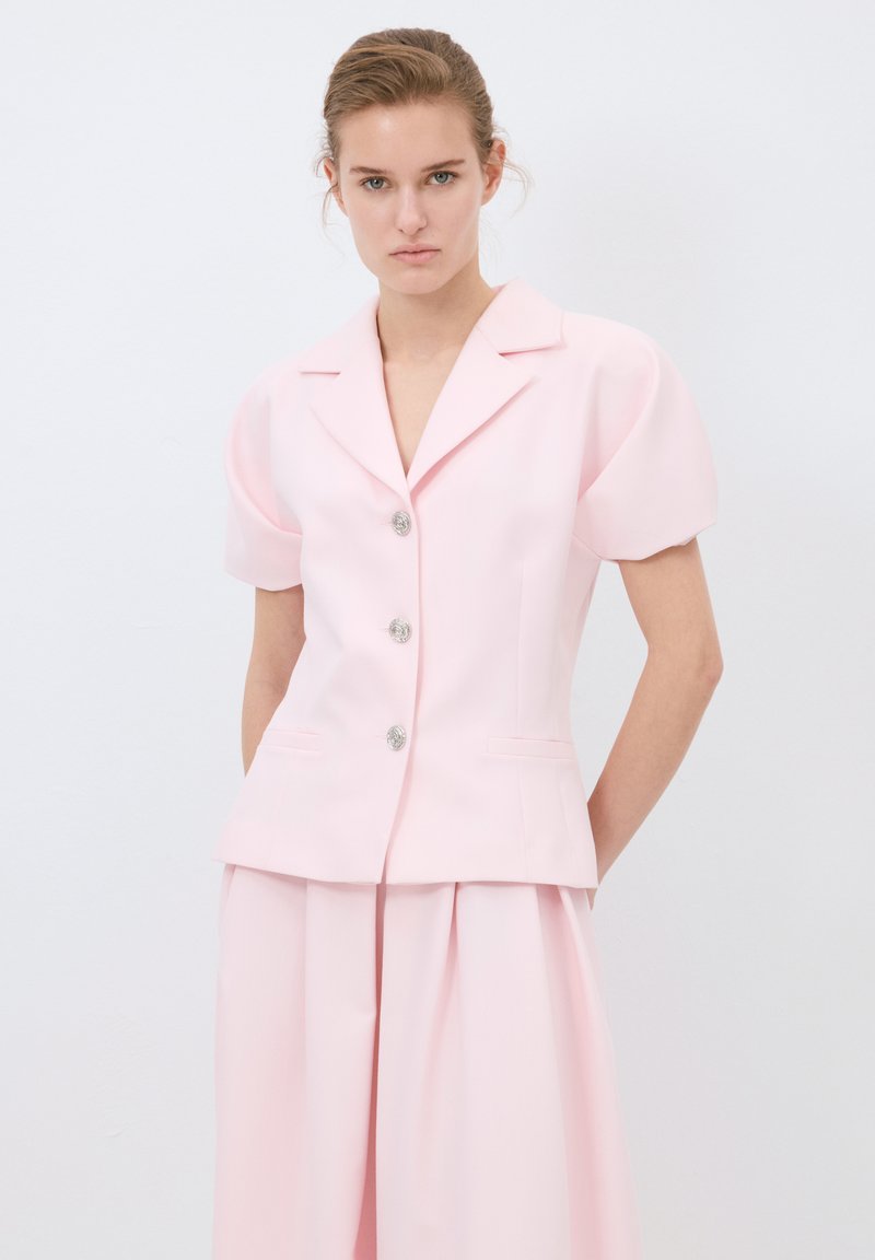 Blazer su misura rosa chiaro con maniche a sbuffo corte, colletto a scalloped, bottoni argentati e tasche laterali, abbinato a pantaloni coordinati.