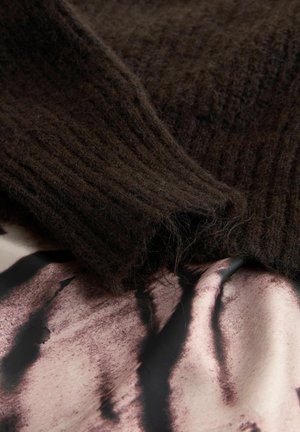 Hnedá rebrovaná pletenina leží na hladkej vzorovanej textílii s abstraktnými tmavými čiarami na svetlom podklade, čím zvýrazňuje kontrast textúr.