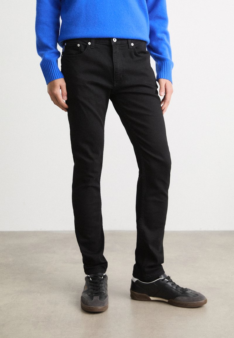 Jeans ajustados negros con bolsillos frontales, cremallera y botón de metal. Combinados con un suéter azul brillante y zapatos deportivos negros.