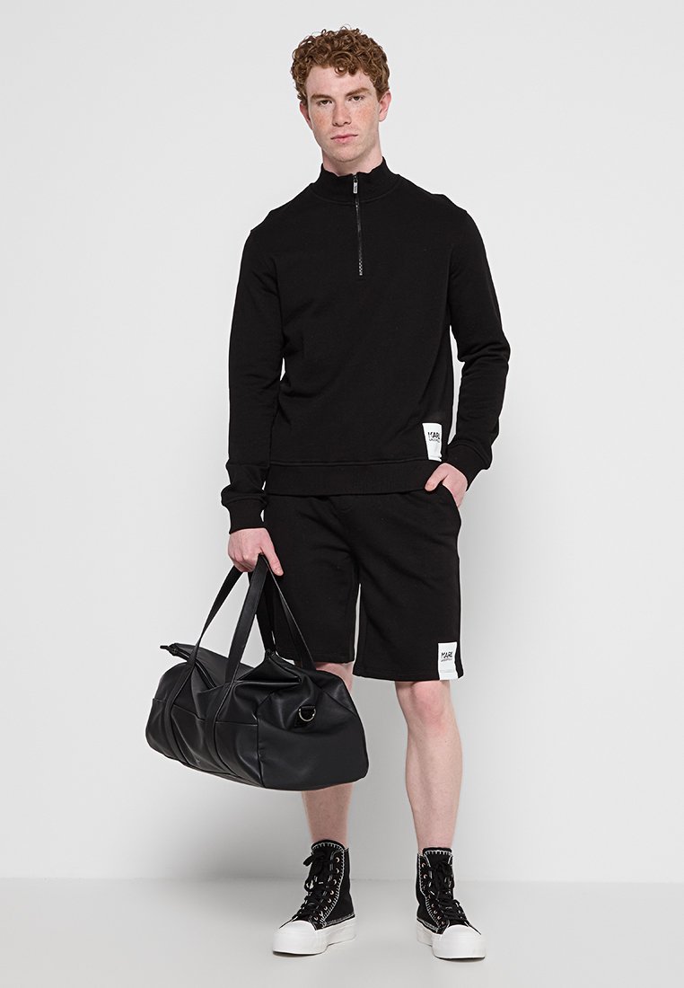 Schwarzer Zip-Hoodie mit gerippten Bündchen, kombiniert mit schwarzen Shorts. Das Modell hält eine große schwarze Tragetasche und trägt schwarze High-Top-Sneaker.
